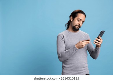 Cheerful young adult does shopping online with his smartphone and card, entering his banking credentials on a website. Person illustrating e commerce, digital payment and modern consumer habits.