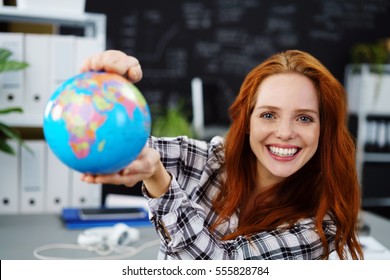 Cheerful Young Adult Female In Red Hair Holding Globe In Classroom. Obscured Chalkboard Behind Her.