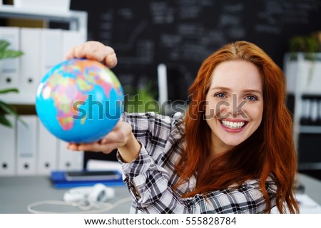Cheerful young adult female in red hair holding globe in classroom. Obscured chalkboard behind her.