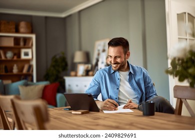 A cheerful young adult man conducting an online interview with a client on a tablet while sitting at a table and holding a pen and a sheet of paper in front of him. - Powered by Shutterstock