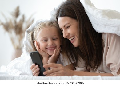 Cheerful young adult mother and cute little child kid daughter bloggers using modern tech smart phone record social media video laughing having fun with mobile apps lying on bed covered with blanket