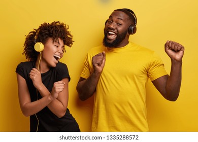 Cheerful young African American bloggers enjoy favourite playlist in headphones, listens audio in music app, feels happiness, dance actively against yellow background, move emotionally, have fun