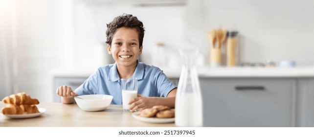 A cheerful young African American boy sits at a kitchen table, smiling as he holds a glass of milk and has a bowl of cookies in front of him, enjoying a delightful snack time. - Powered by Shutterstock