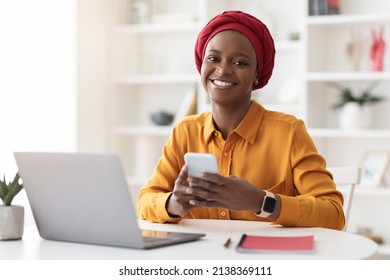 Cheerful young african american businesswoman in red turban sitting at workdesk at cozy office, using modern smartphone and laptop, chatting with clients or using business app, copy space - Powered by Shutterstock