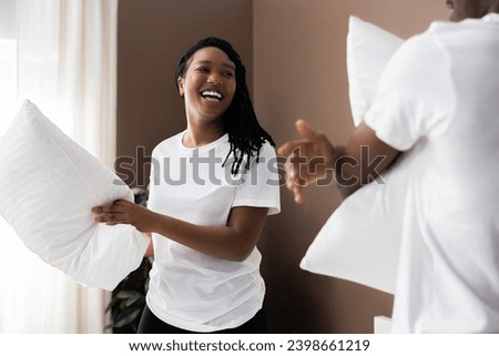 Cheerful young African American dark skinned couple having fun pillow fight in bedroom with husband.