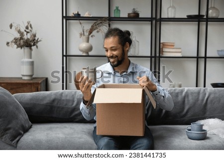 Cheerful young African American homeowner man unpacking cardboard box, moving into new home. Customer guy opening parcel with dish, producing cups, ceramic mugs from carton container on couch