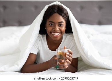 Cheerful young african american lady laying under blanket and enjoying aromatic herbal tea in bed, smiling at camera, morning with healthy green tea, free space, bedroom interior, detox concept