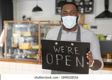 A cheerful young African American male waiter wearing mask holding in hands open sign indoors in own bakery shop. Reopening bakehouse after lockdown, pandemic, epidemic