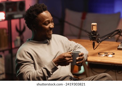 Cheerful young African American male blogger drinking coffee during live podcast in recording studio - Powered by Shutterstock