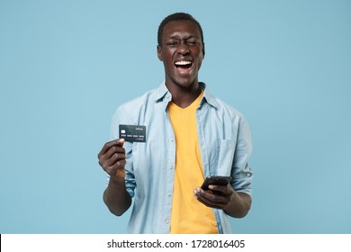 Cheerful Young African American Man Guy In Casual Shirt, Yellow T-shirt Posing Isolated On Blue Wall Background. People Lifestyle Concept. Mock Up Copy Space. Using Mobile Phone Hold Credit Bank Card