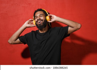Cheerful young african american man with dreadlocks 20s wearing black casual t-shirt posing listen music with headphones looking aside isolated on bright red color wall background studio portrait