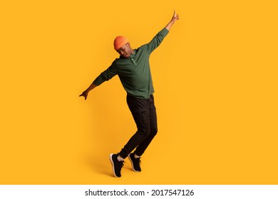 Cheerful young african american man in orange hat standing on tiptoes over yellow background, full length shot of happy millennial black hipster guy dancing and having fun in studio, copy space
