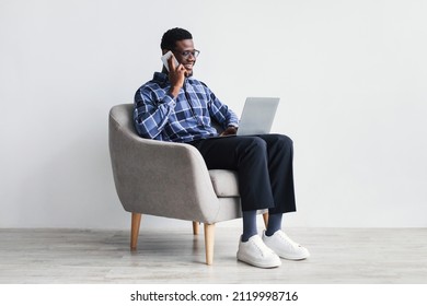 Cheerful Young African American Man With Laptop Pc Communicating On Cellphone, Sitting In Armchair Against White Studio Wall. Millennial Black Man Speaking To Friend, Using Portable Computer