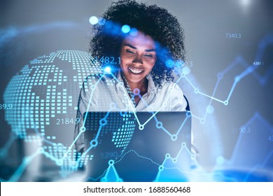 Cheerful Young African American Woman Trader Using Laptop In Blurry Office With Double Exposure Of Blurry Financial Chart And Planet Hologram. Concept Of Stock Market And Technology. Toned Image