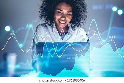 Cheerful young African American woman using laptop at blurry table with double exposure of financial graph. Concept of trading and financial success. Toned image