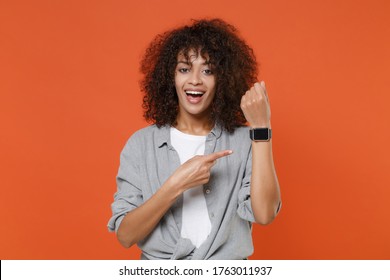 Cheerful young african american woman in gray casual clothes isolated on orange background studio portrait. People lifestyle concept. Mock up copy space. Pointing index finger on smart watch on hand