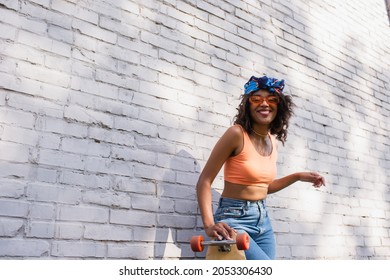 cheerful young african american woman in sunglasses and headscarf holding longboard near brick wall