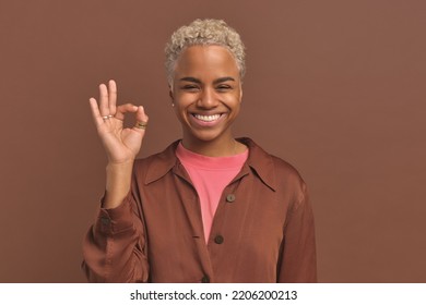 Cheerful Young African American Woman Student Shows OK Gesture And Smiles When Answering Question Or Confirming Correctness Of Action Stands In Monochromatic Brown Studio. Yes, I Agree Concept