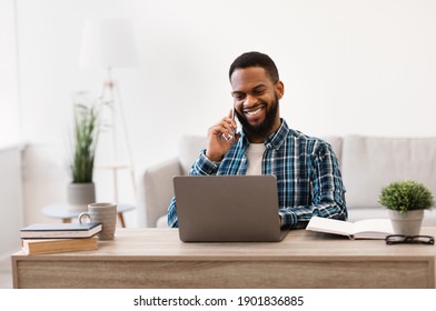 Cheerful Young African Businessman Talking On Cellphone Sitting At Laptop Working In Modern Office Indoor. Mobile Communication And Distant Work, Male Entrepreneurship Concept