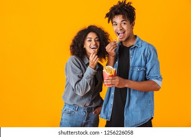 Cheerful young african couple standing isolated over orange background, eating french fries