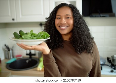 Cheerful Young African Ethnicity Woman With Long Black Curly Hair Standing At Kitchen Holding Plate With Greens And Cucumber, Preparing Dinner Or Lunch. Healthy Food, Lifestyle. Key To Longevity