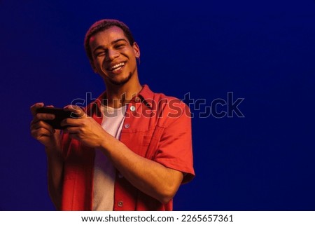 Cheerful young african guy playing online game on mobile phone isolated over blue neon studio wall