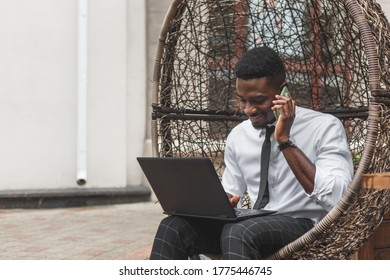 Cheerful young African man in formal wear talking on the mobile phone and working on laptop while sitting on egg chair at outdoors cafe