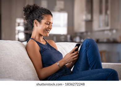 Cheerful Young African Woman Using Smartphone While Sitting On Couch. Black Smiling Woman Using App On Cellphone At Home. Beautiful Casual Girl Relaxing While Chatting On Mobile Phone. 