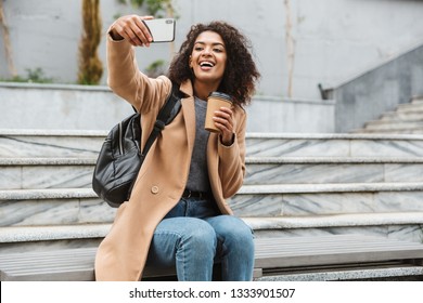 Cheerful Young African Woman Wearing Coat Sitting Outdoors, Holding Takeaway Coffee Cup, Taking A Selfie