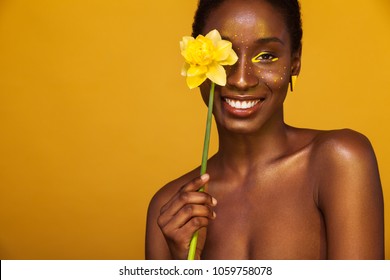 Cheerful young african woman with yellow makeup on her eyes. Female model laughing against yellow background with yellow flower.