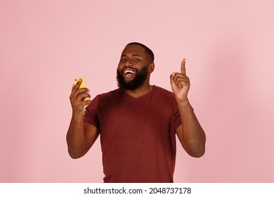 Cheerful young African-American man in t-shirt with mobile phone gets genius idea on light pink background in studio closeup - Shutterstock ID 2048737178