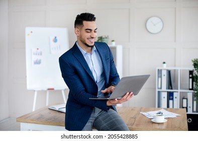 Cheerful young Arab businessman in formal suit using laptop computer, working online at modern office. Young male CEO conducting meeting or conference with employees on web