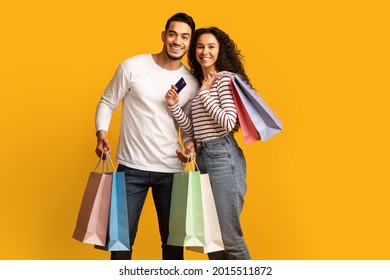 Cheerful Young Arab Couple Posing With Credit Card And Bright Shopping Bags, Happy Middle Eastern Man And Woman Making Purchases Together, Standing Isolated Over Yellow Background, Copy Space