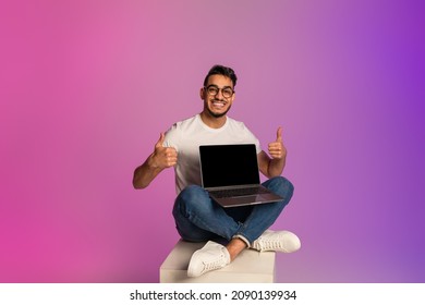 Cheerful Young Arab Guy Holding Laptop Pc With Empty Screen, Showing Thumb Up Gesture, Recommending New Website, Advertising Your Product In Neon Light. Mockup For Design