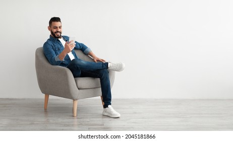 Cheerful young Arab man having online business meeting on smartphone, sitting in cozy armchair near white wall, banner design with free space. Eastern guy communicating remotely on mobile device