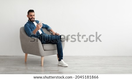 Cheerful young Arab man having online business meeting on smartphone, sitting in cozy armchair near white wall, banner design with free space. Eastern guy communicating remotely on mobile device
