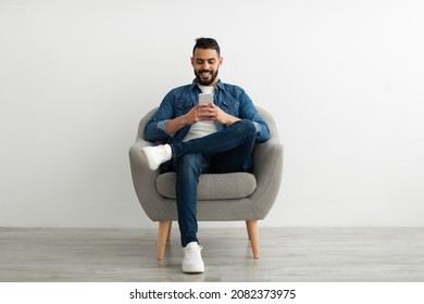 Cheerful young Arab man using smartphone, having online chat, sitting in armchair, browsing social media against white studio wall, blank space. Remote communication, blogging concept