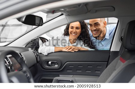 Cheerful young arab man and woman buying new car, checking automobile salon through the window. Smiling middle-eastern couple looking inside beautiful black auto in showroom, purchasing auto