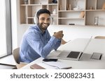 Cheerful young Arab online student guy portrait. Happy businessman, freelancer, influencer, support center employee in headphones sitting at workplace table with laptop, looking at camera, smiling