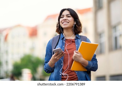 Cheerful young arab student female listening music with smartphone and earphones outdoors, smiling middle eastern woman carrying workbooks and backpack while walking on city street, copy space - Shutterstock ID 2206791389