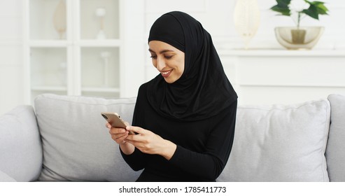 Cheerful Young Arabian Beautiful Woman In Black Hijab Sitting On Couch And Texting Message On Smartphone At Home. Pretty Smiled Female Muslim In Headscarf Tapping And Scrolling On Mobile Phone.