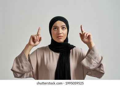 Cheerful young arabic woman in hijab pointing fingers up while looking upstairs on light background. Beautiful muslim lady