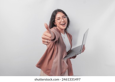 Cheerful young Asian business woman showing thumbs up and holding laptop on isolated white background.