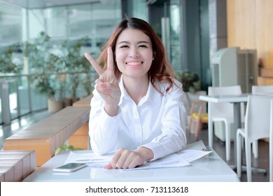 Cheerful young Asian business woman holding two finger as sign as fighting with job at office.