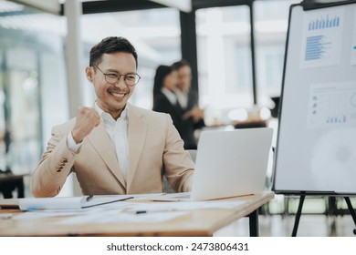 Cheerful young Asian businessman smiling in bright modern office working on laptop computer checking charts, graphs and paper. - Powered by Shutterstock