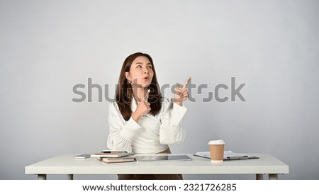 A cheerful young Asian businesswoman or female office worker looking up and pointing her fingers up at empty space, a surprised gesture, a wow, special deal, or hot promotion facial expression.