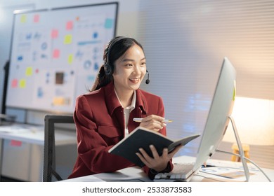 Cheerful young asian businesswoman works late at night in a modern office, taking notes on her computer with a headset, showing determination and confidence - Powered by Shutterstock