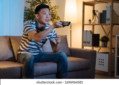 Cheerful Young Asian Chinese Man Watching Movies On Tv At Home Late In Evening. Handsome Guy Relax Sitting In Comfort Couch Stay Up Late At Night Enjoy Television Program. Male Holding Remote Switch.