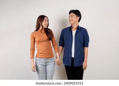 Cheerful young Asian couple in casual outfits smiling and looking at each other with holding hands. Man and woman being in love