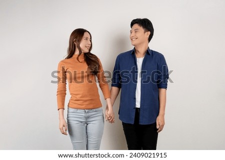 Cheerful young Asian couple in casual outfits smiling and looking at each other with holding hands. Man and woman being in love